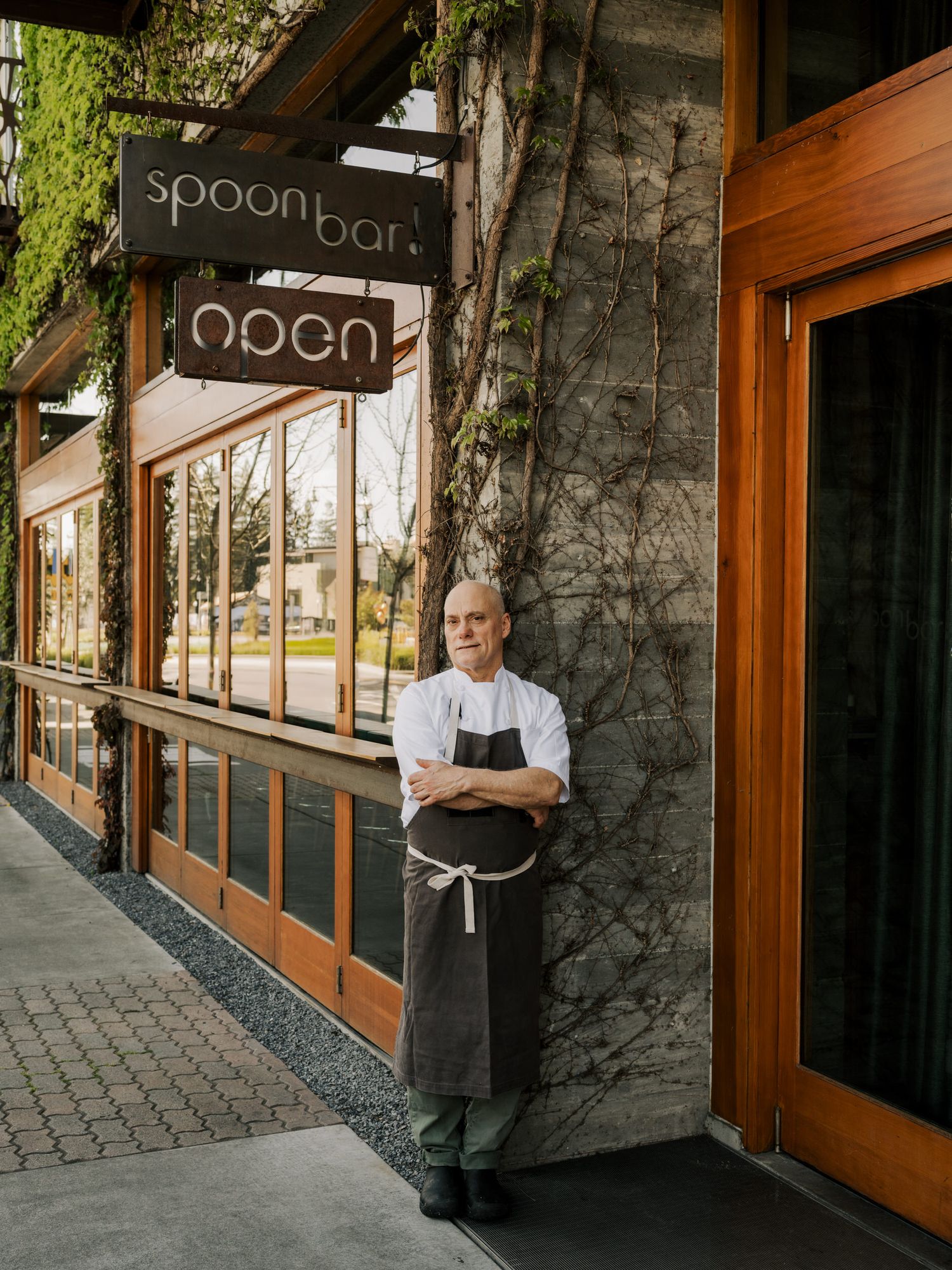 Chef in white coat and black apron stands outside Spoonbar restaurant entrance with open sign and stone exterior.