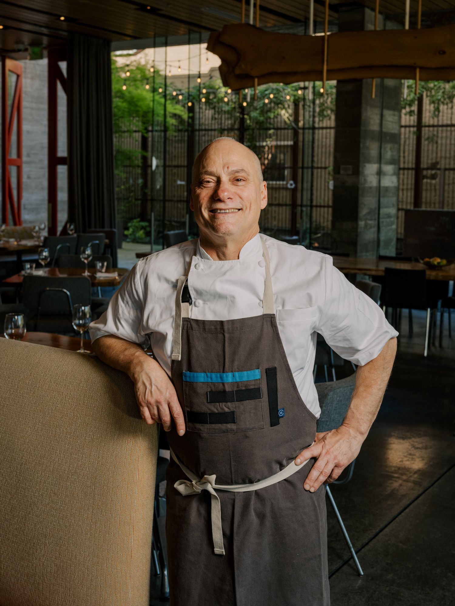 A professional in a dark gray apron and white shirt poses in a sophisticated indoor setting.