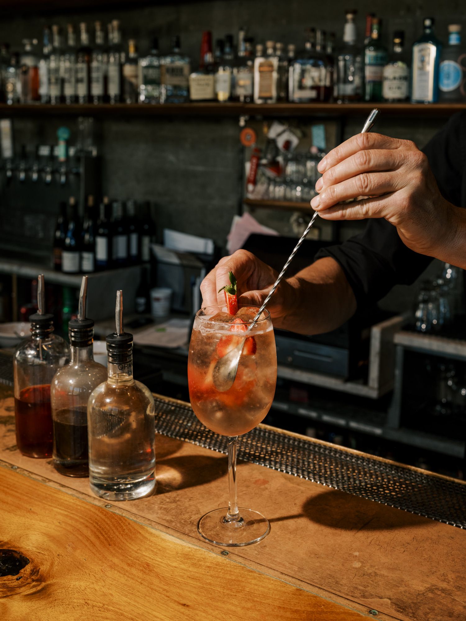 A hand stirs a cocktail with a metal spoon in a dimly lit bar setting, showcasing various bottles in the background.