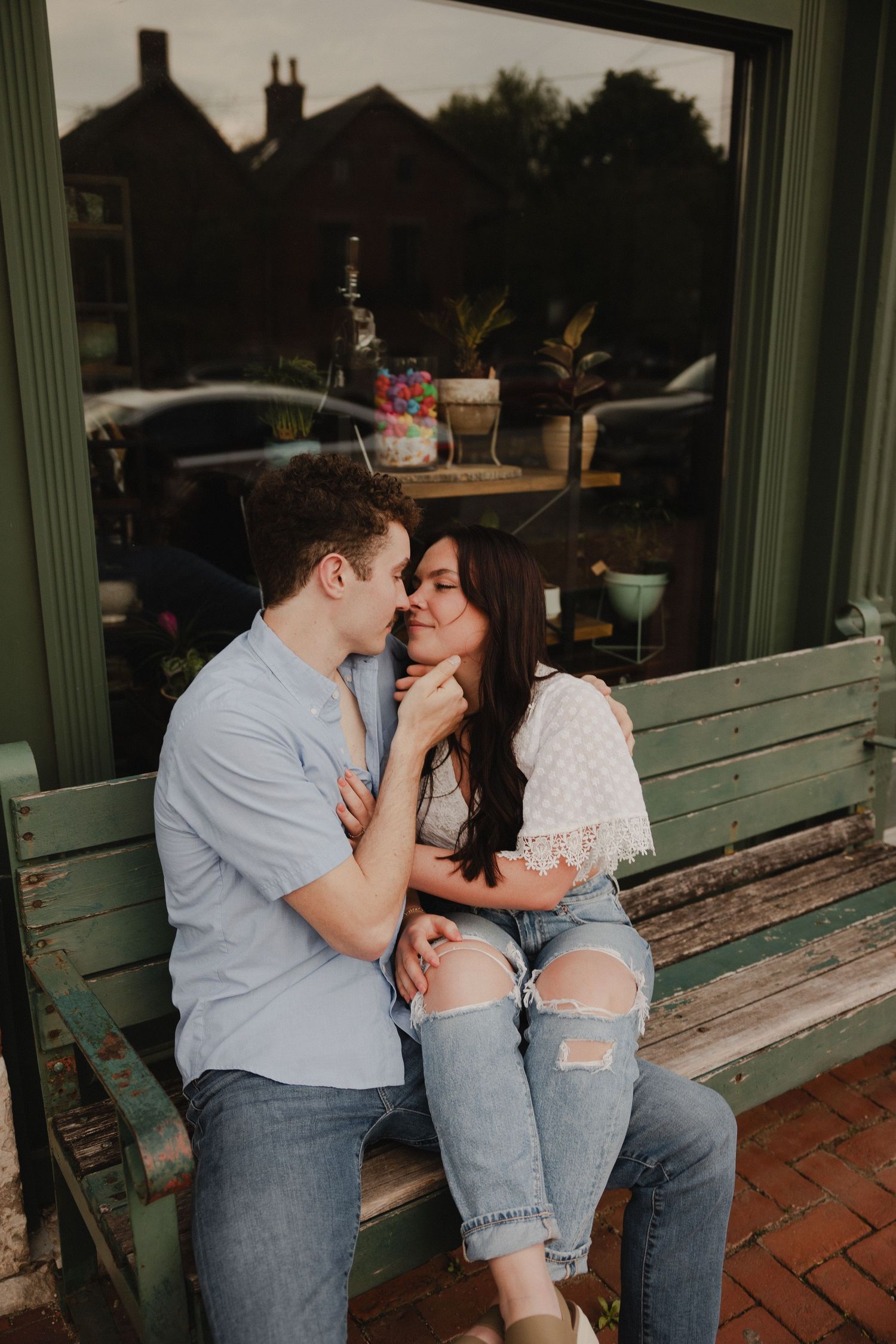 A romantic couple shares an intimate moment while sitting on a green wooden bench near a window.