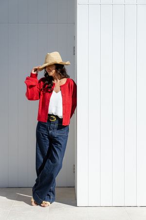 A person in a red cardigan and wide-leg jeans leans against a white wall while wearing a straw sun hat.