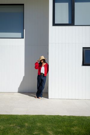 A figure in a red jacket and straw hat leans against a white modern building with geometric windows on a sunny day.