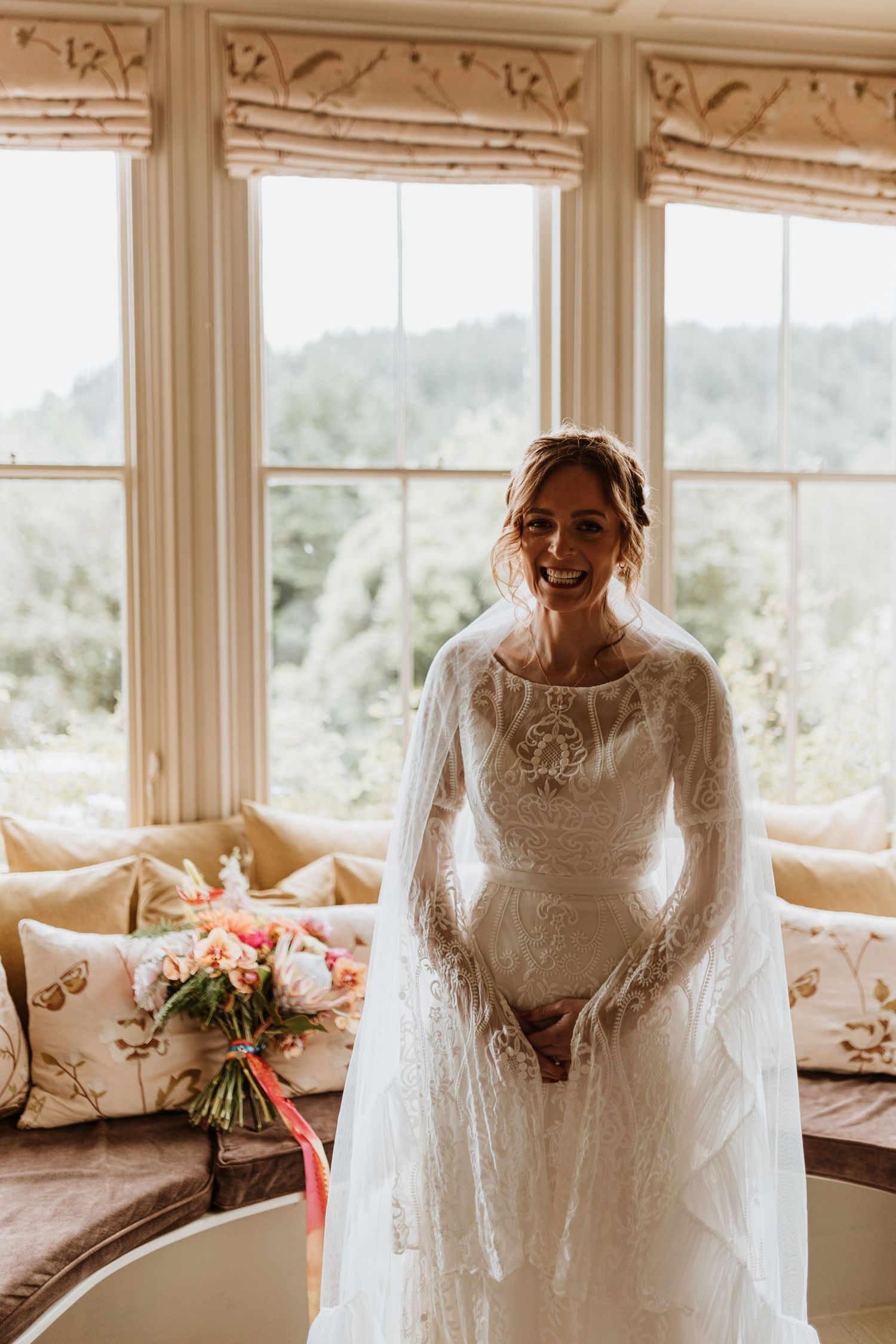 A bride in a long sleeve lace wedding dress stands near sunny windows, the veil draping elegantly behind her.