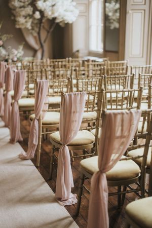 Gold chairs with blush pink fabric sashes line up at an elegant indoor wedding ceremony venue at Rise Hall.