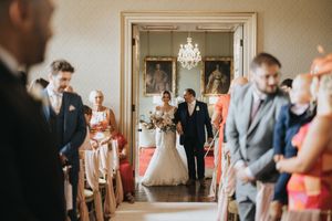 A wedding ceremony in progress with guests seated in a grand room with chandelier as the couple enters through doorway at Rise Hall.