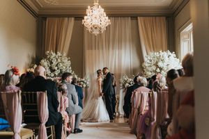 Elegant indoor wedding ceremony in ballroom with crystal chandelier and tall white floral arrangements along the aisle at Rise Hall.