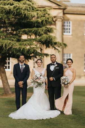Wedding party poses on a manicured lawn in front of an elegant stone building with a cypress tree framing the scene at Rise Hall.