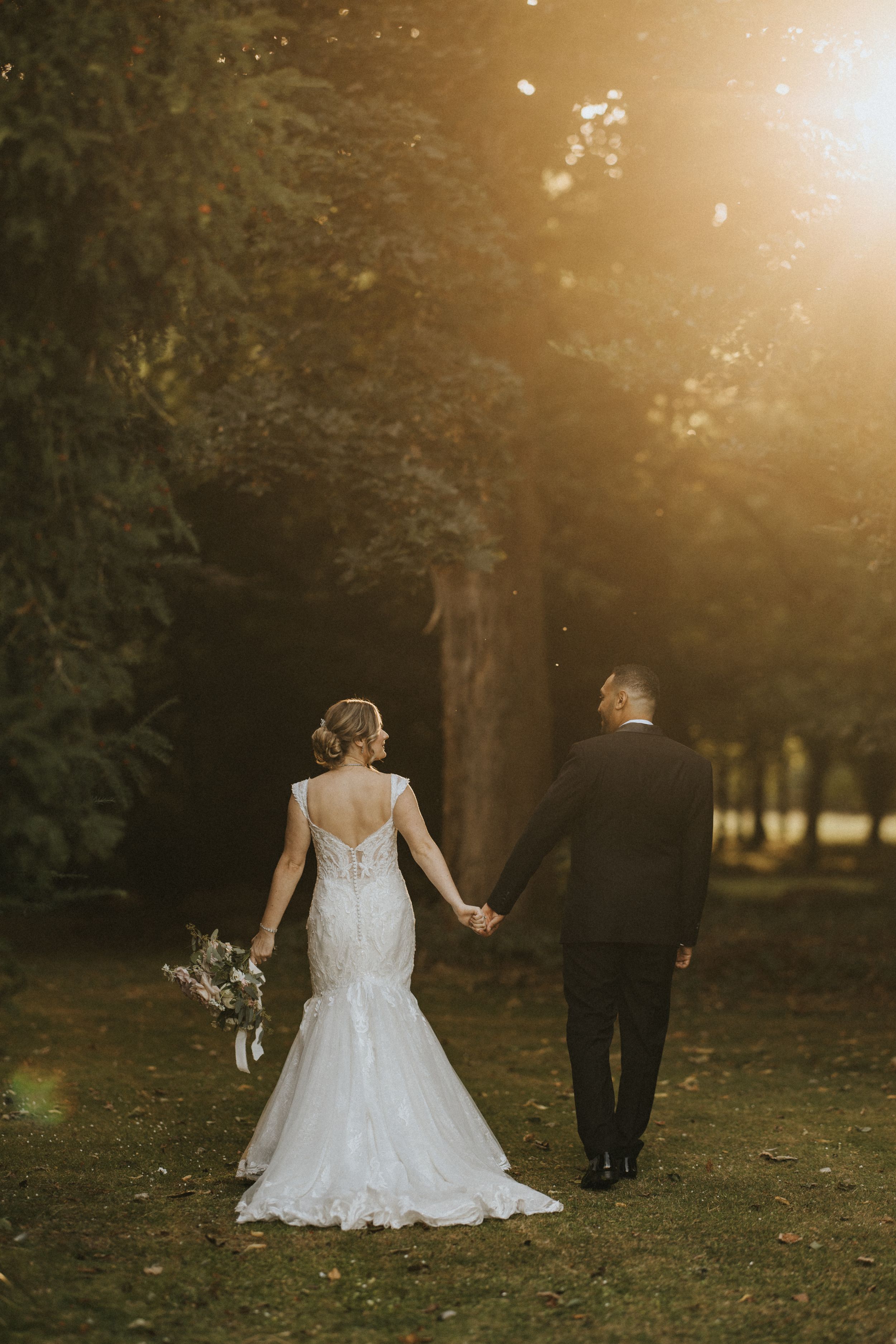 A bride in a lace mermaid gown and groom holding hands walk away into a sunlit wooded area during sunset at Rise Hall.