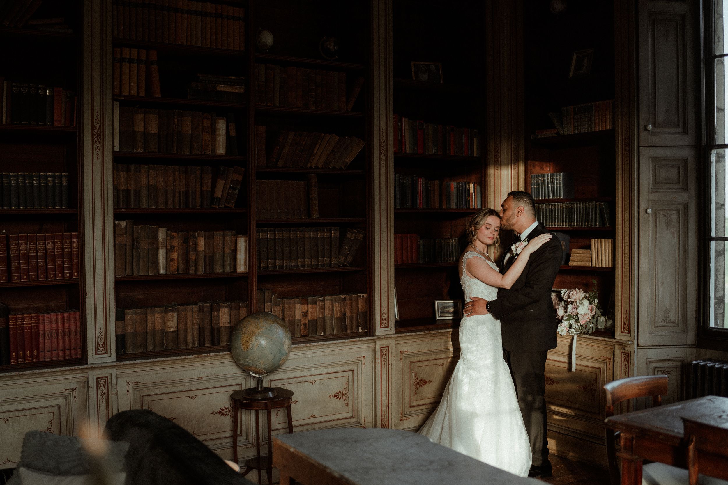 A couple shares an intimate moment in an elegant library setting with vintage books and a globe on display at Rise Hall.