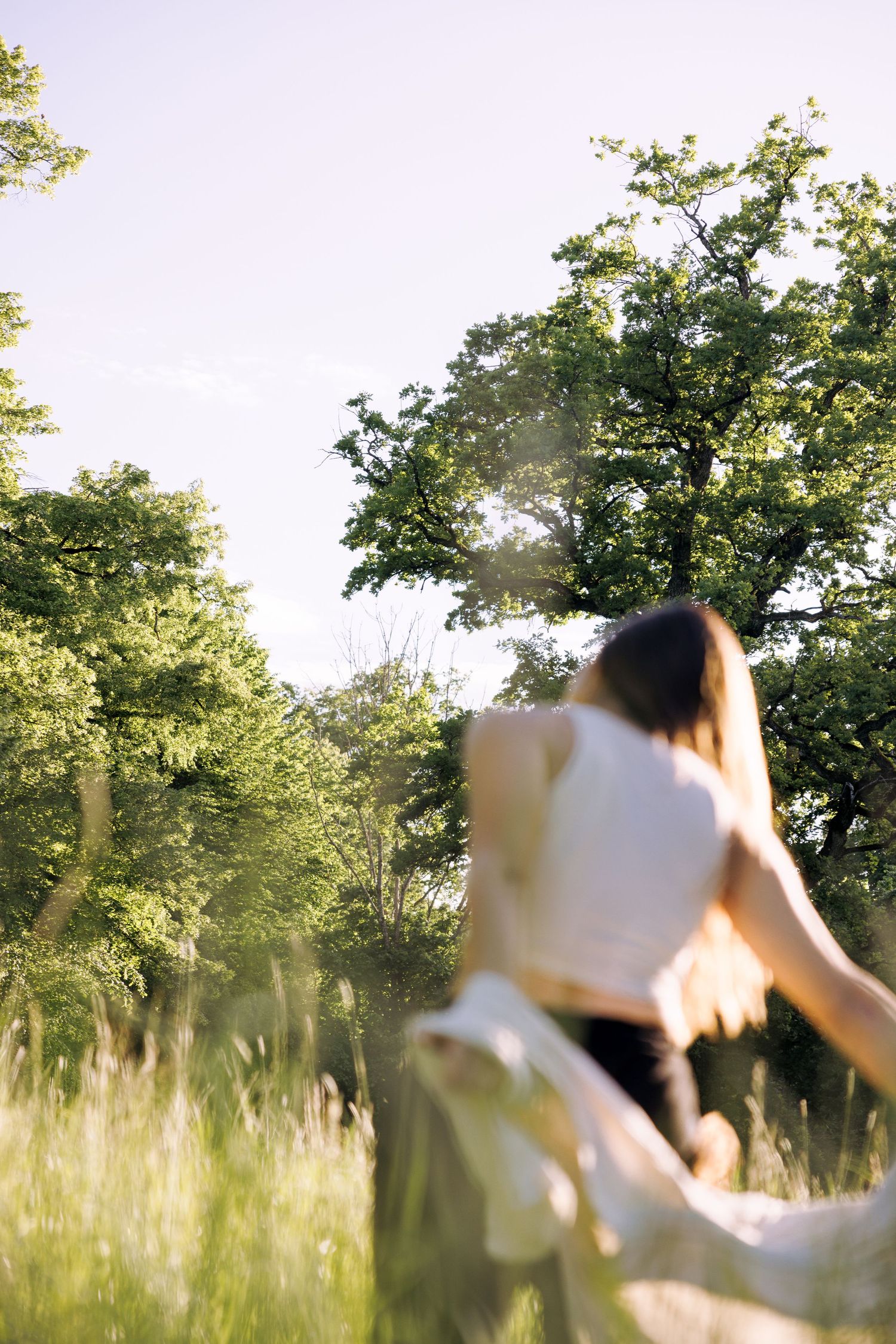 Person in white top twirling joyfully in a sunlit meadow with trees in background.