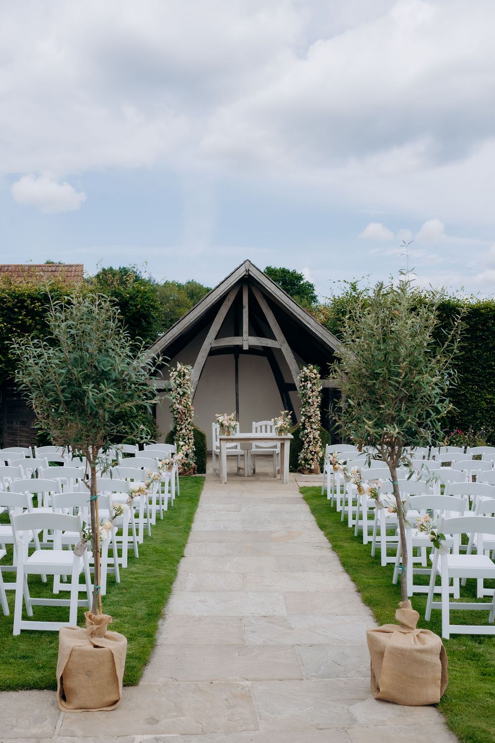 White wedding chairs line a stone path leading to a modern chapel structure under cloudy skies at an outdoor ceremony venue.