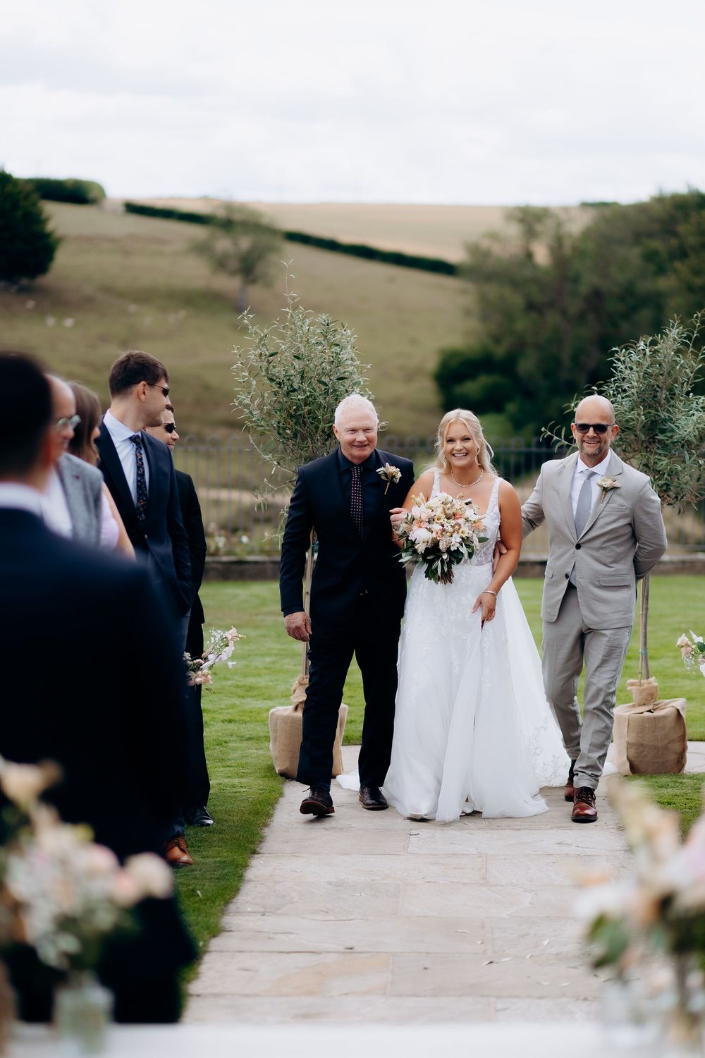 Wedding party walking down an outdoor garden path with rolling hills in the background on a cloudy day.
