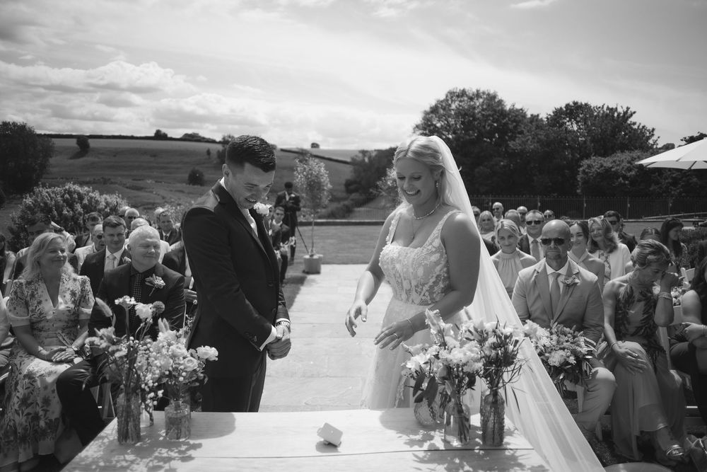 Black and white photograph of an outdoor wedding ceremony with guests seated on both sides while couple exchanges vows.