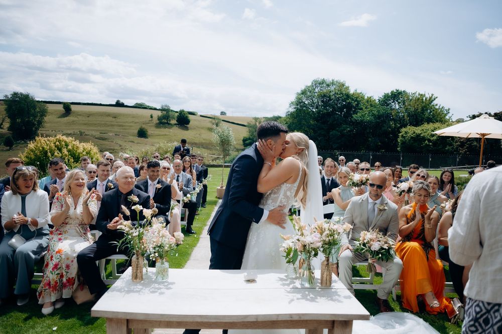A scenic outdoor wedding ceremony takes place on a hilltop with guests seated on chairs and floral arrangements on display.