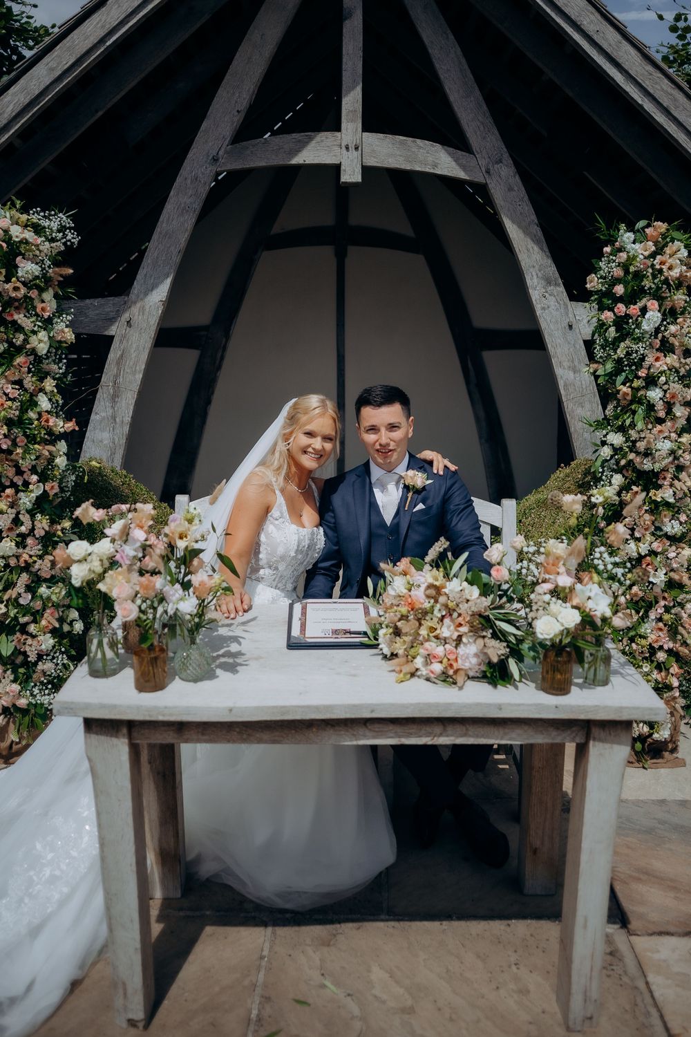 A couple signs their wedding ceremony paperwork at a rustic altar adorned with pink and white floral arrangements.