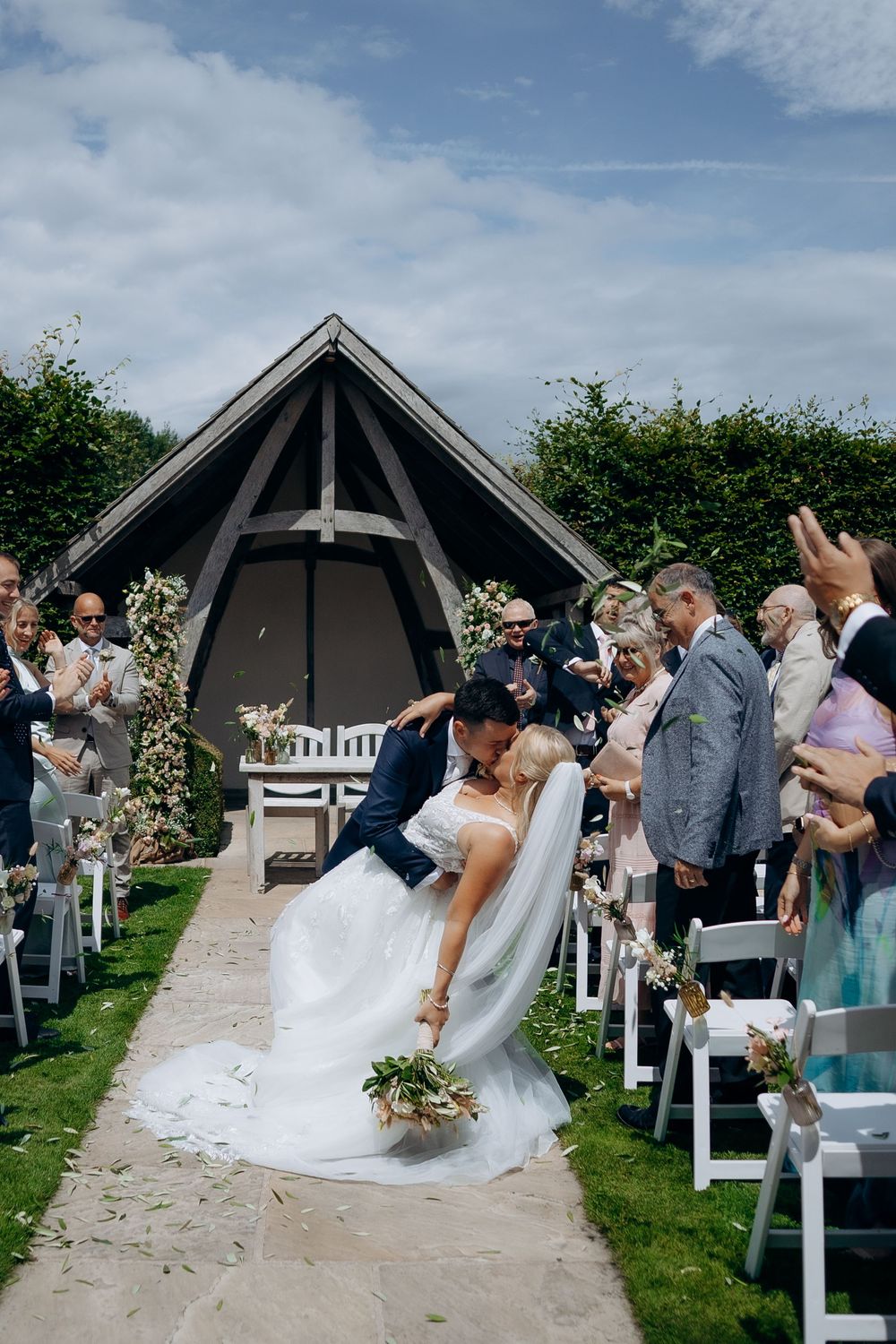 A romantic outdoor wedding ceremony takes place at a garden venue with a rustic wooden gazebo and white chairs along the aisle.