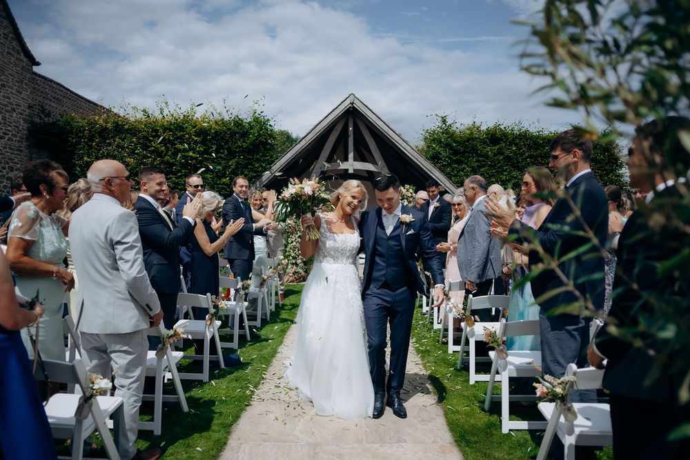 Guests gather for an outdoor wedding ceremony in a lush garden setting with white chairs arranged in rows.