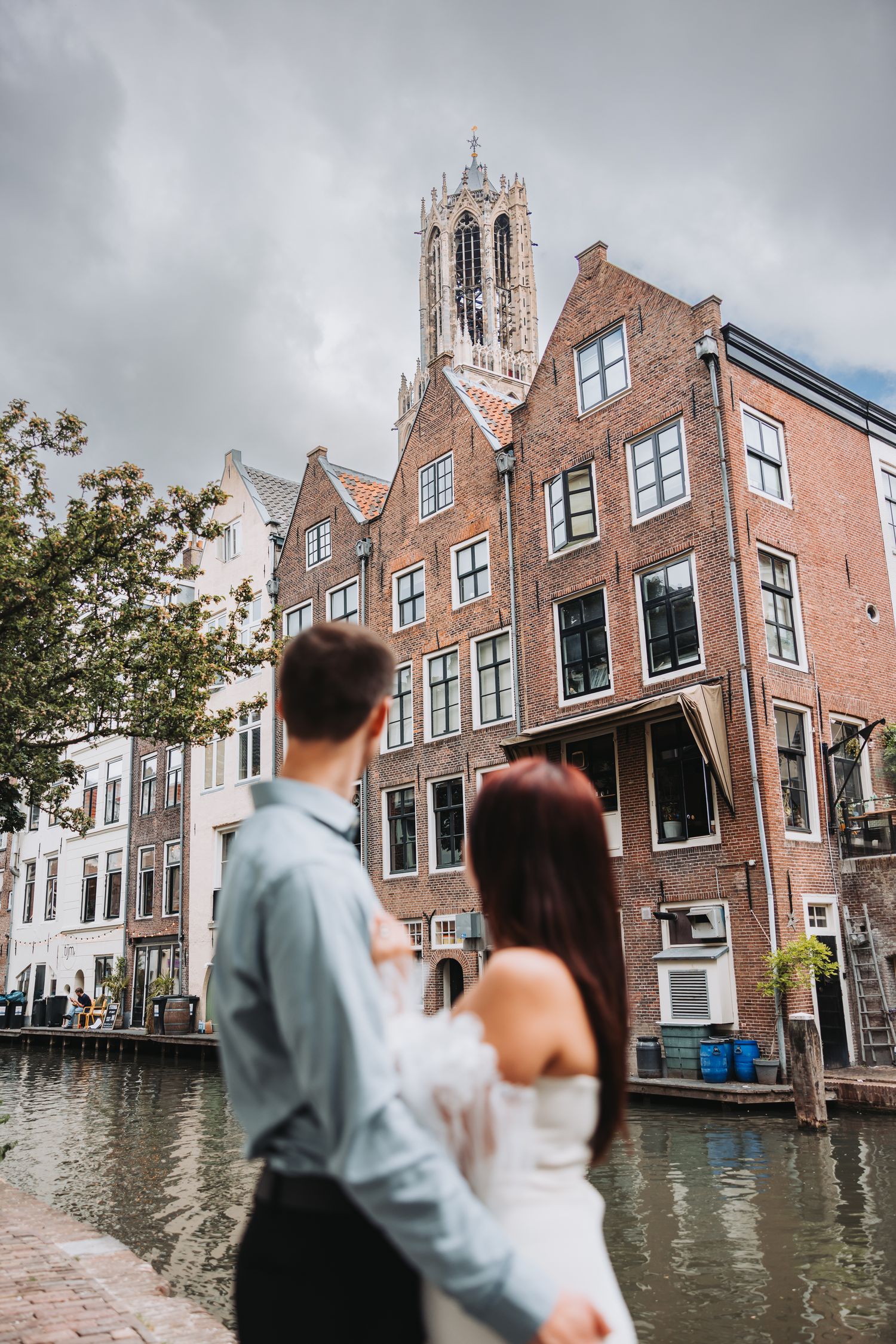 A bridal couple admires the historic architecture and canal views in Utrecht, Netherlands with a Gothic church tower in the background.