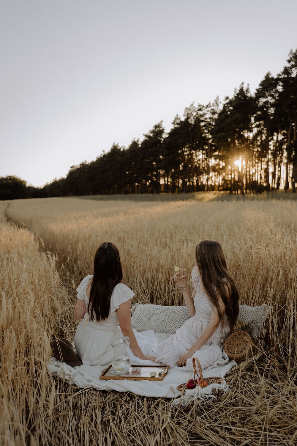 Two people in white dresses having a picnic in a golden wheat field at sunset with trees in the background.