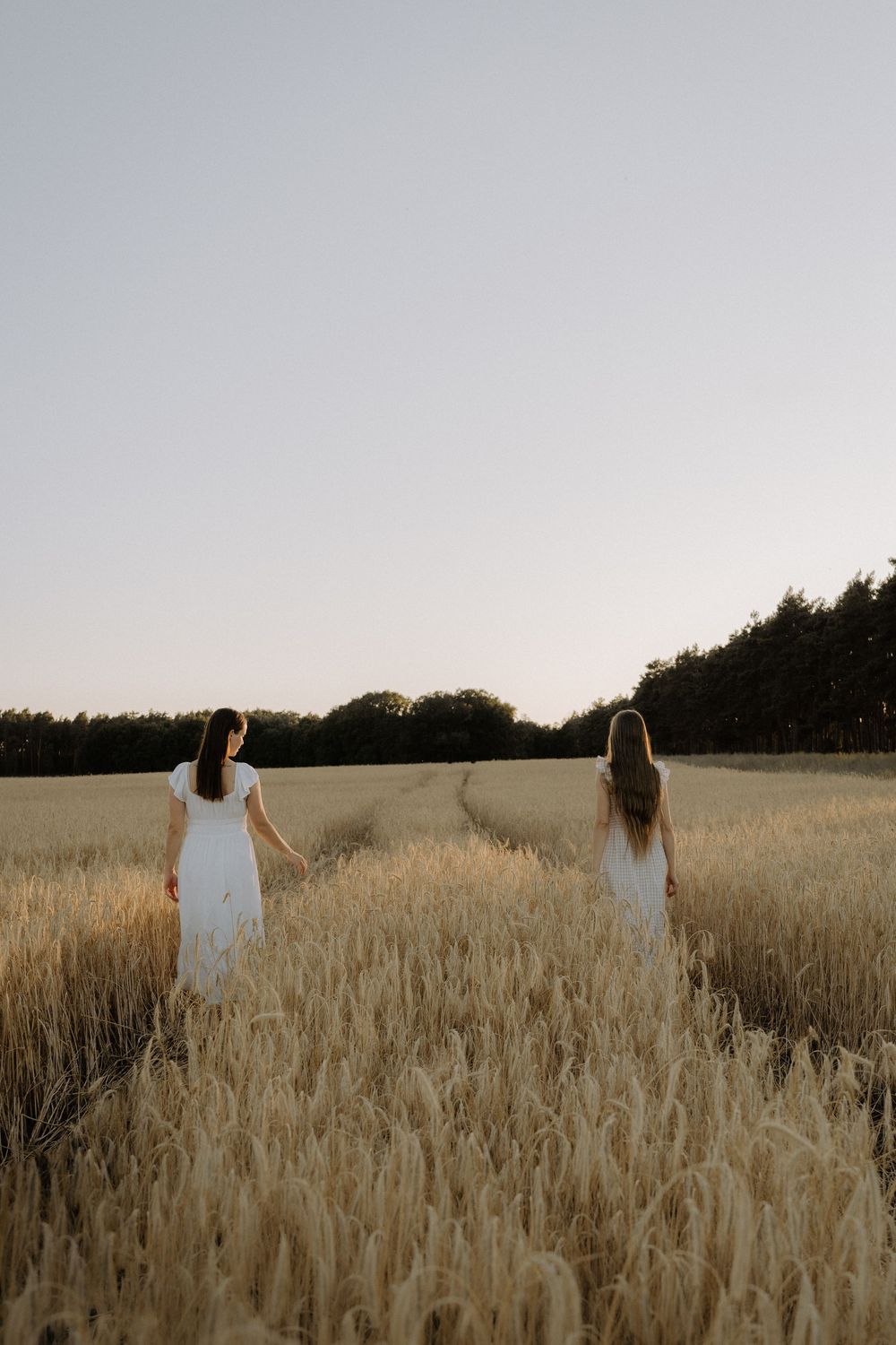 Two women in white dresses stand together in a golden wheat field at sunset with trees in the distance.