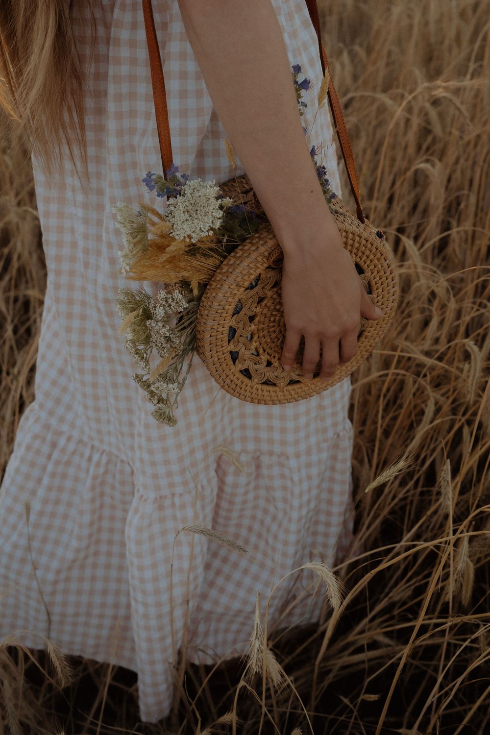 Person in gingham dress holds woven rattan bag with dried flowers while standing in tall grass field at sunset.