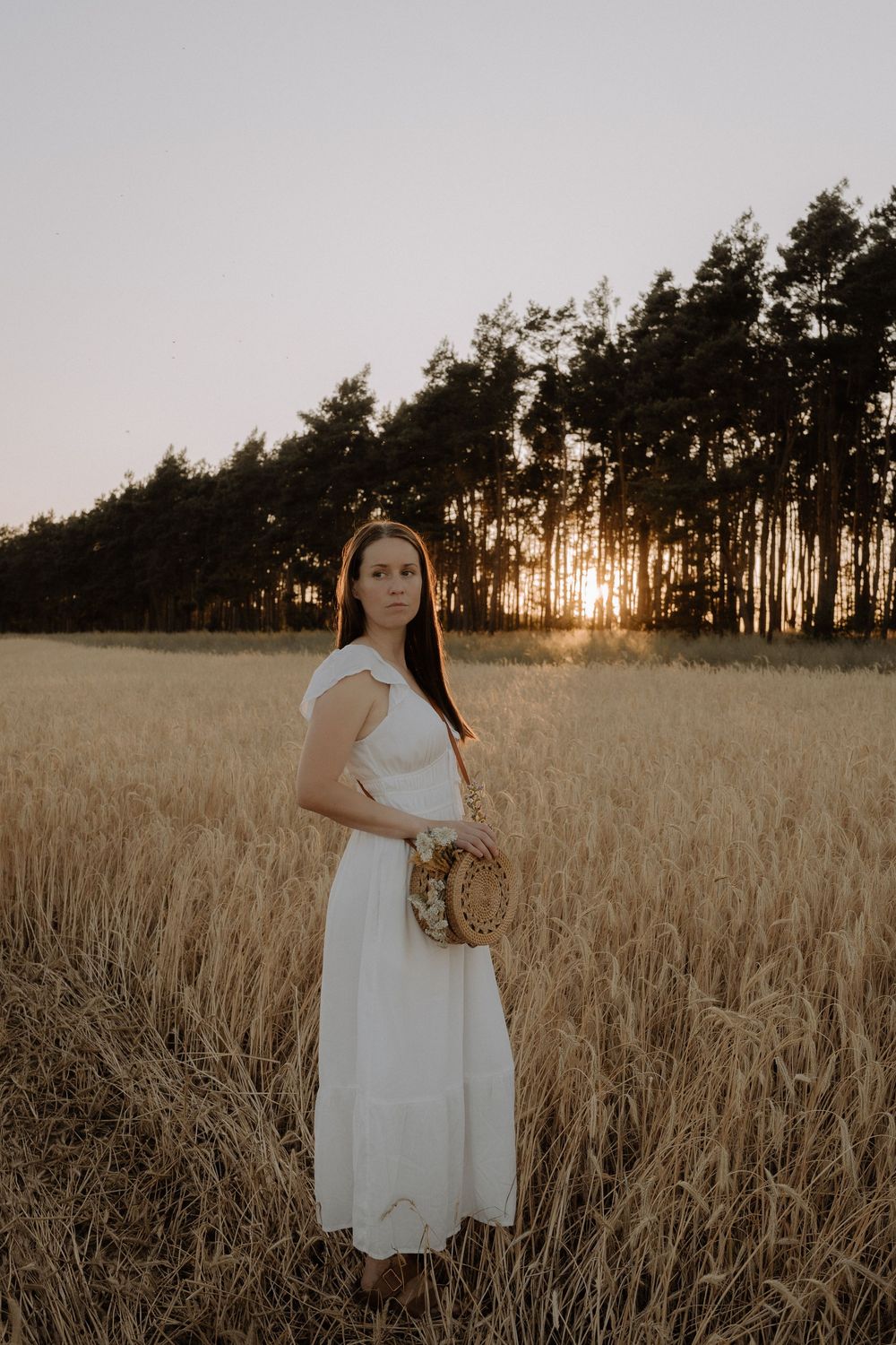 A white dress flows in a golden wheat field at sunset with silhouetted pine trees in the background.
