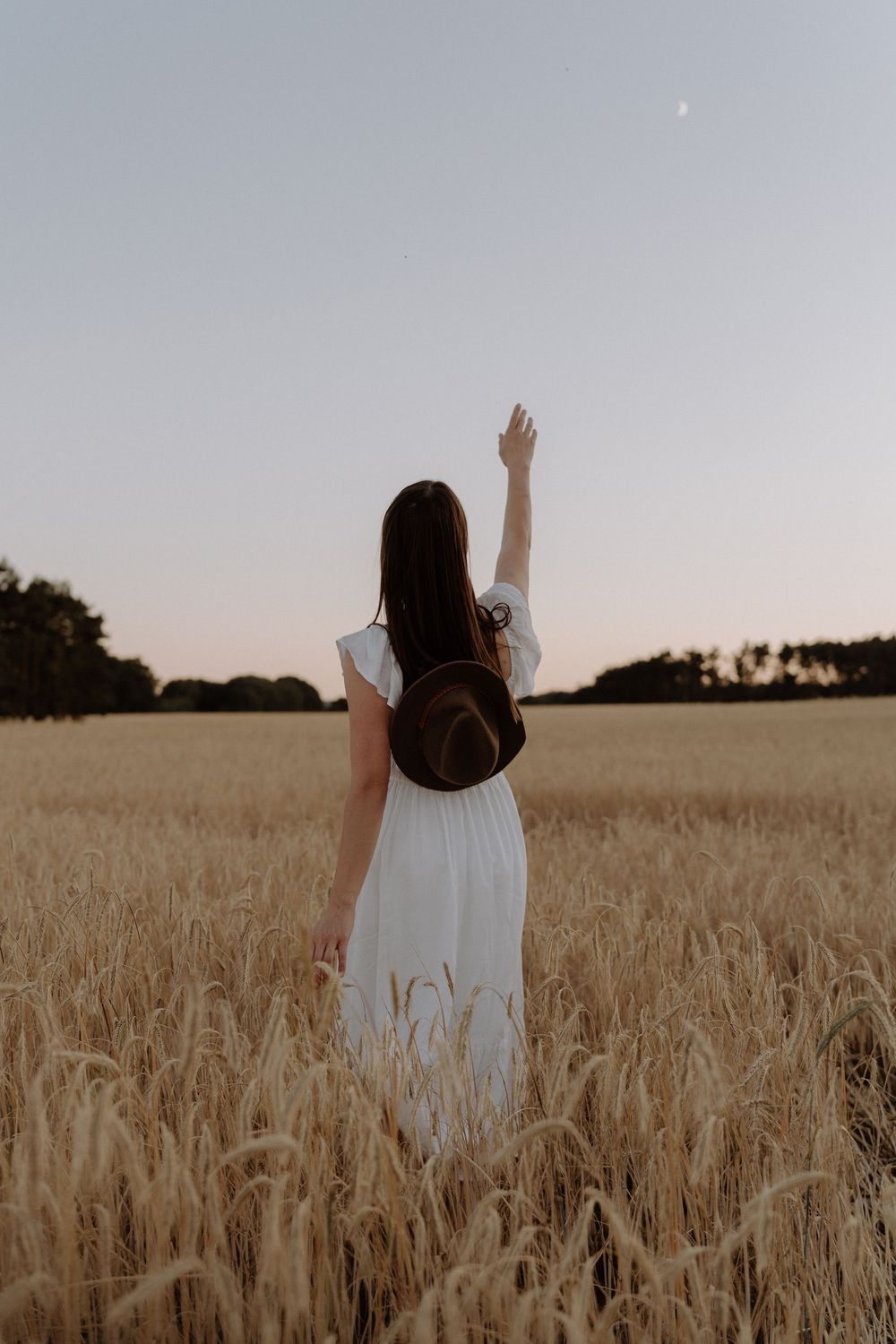A person in a white dress stands with raised arms in a golden wheat field at sunset.