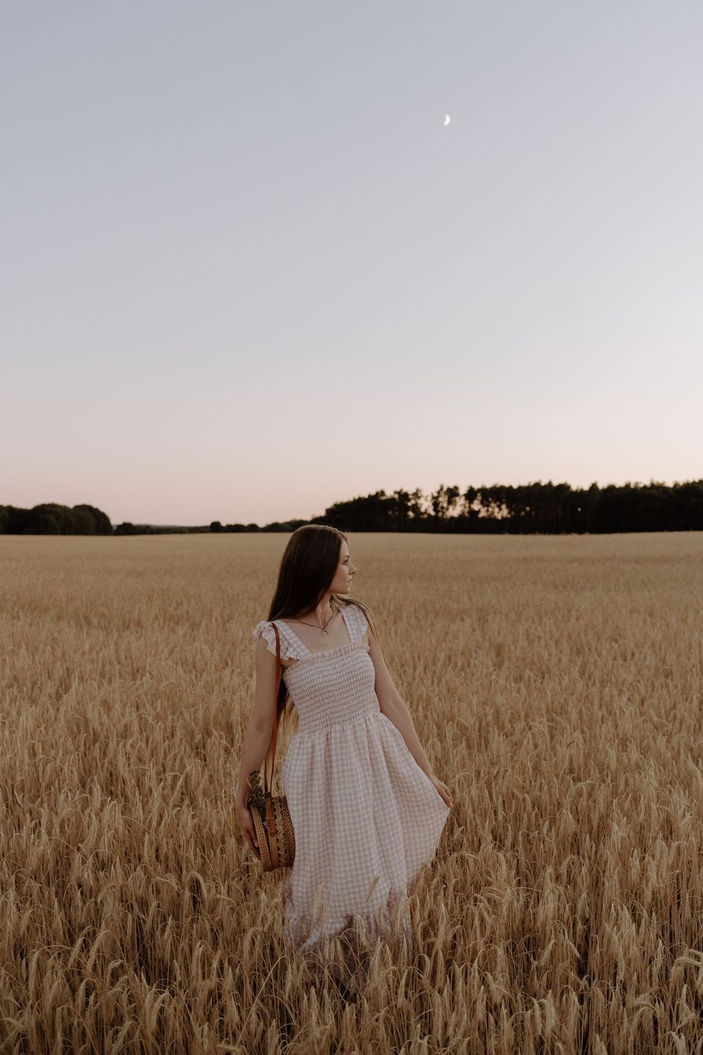 A person in a white gingham dress stands among golden wheat stalks at sunset with trees silhouetted in the distance.