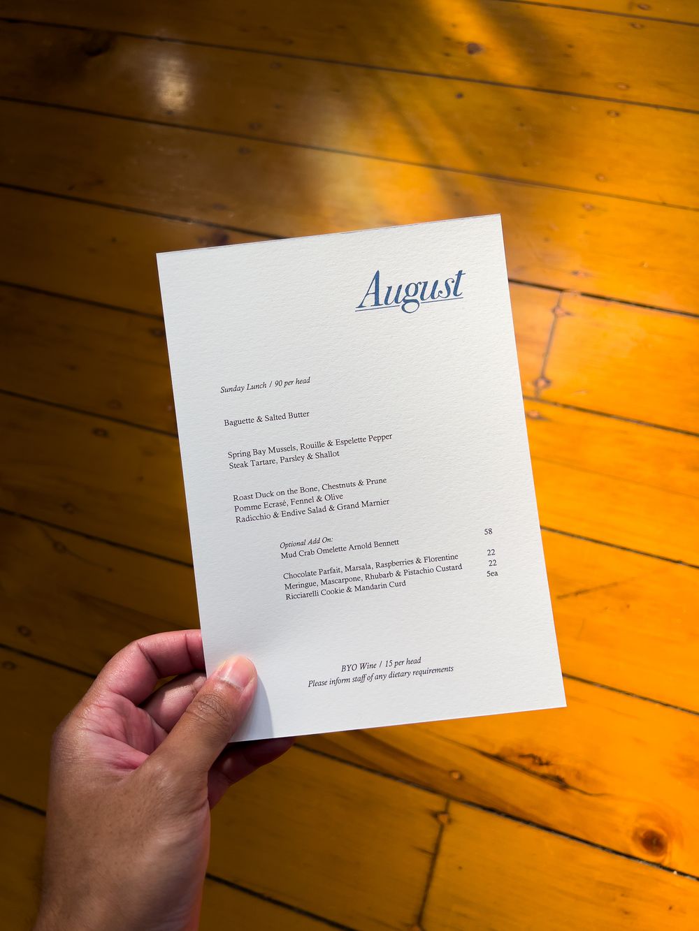 A rustic wooden table is illuminated with warm orange light as a hand holds a menu titled 'August' in an elegant font.