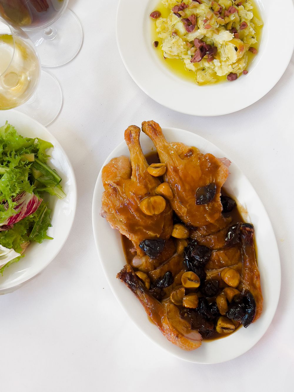 Roasted chicken with prunes and garlic served alongside a fresh green salad and white wine on a white table setting.
