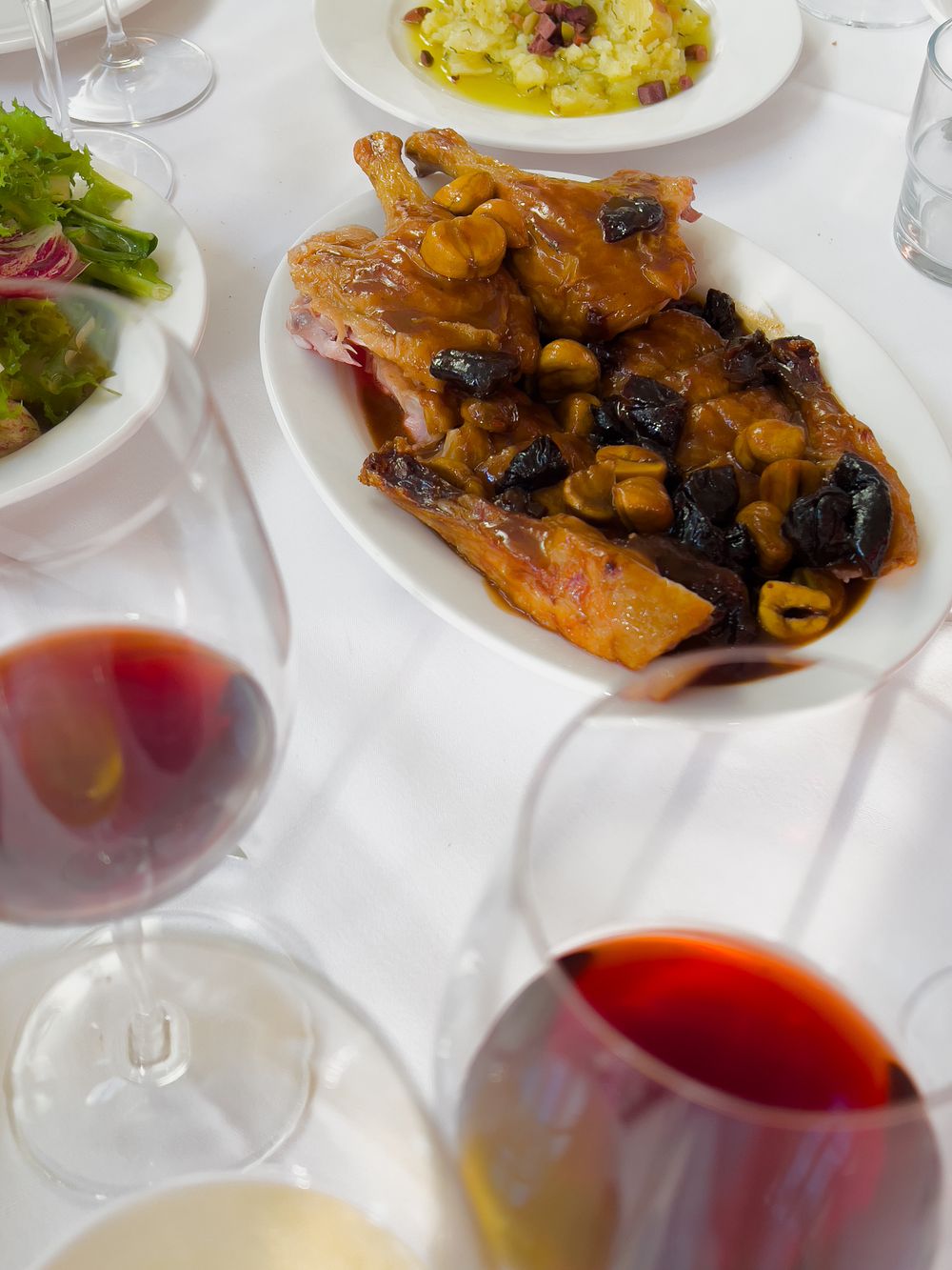 Roasted meat with prunes in sauce served on white plates alongside glasses of red wine at a formal dining setting.