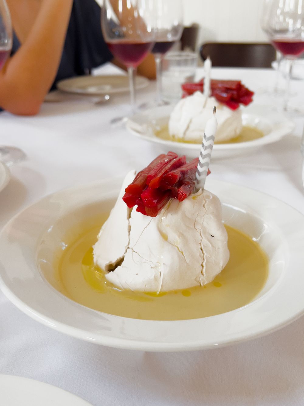 A white meringue dessert drizzled with olive oil and garnished with red flower petals on a pristine white plate.