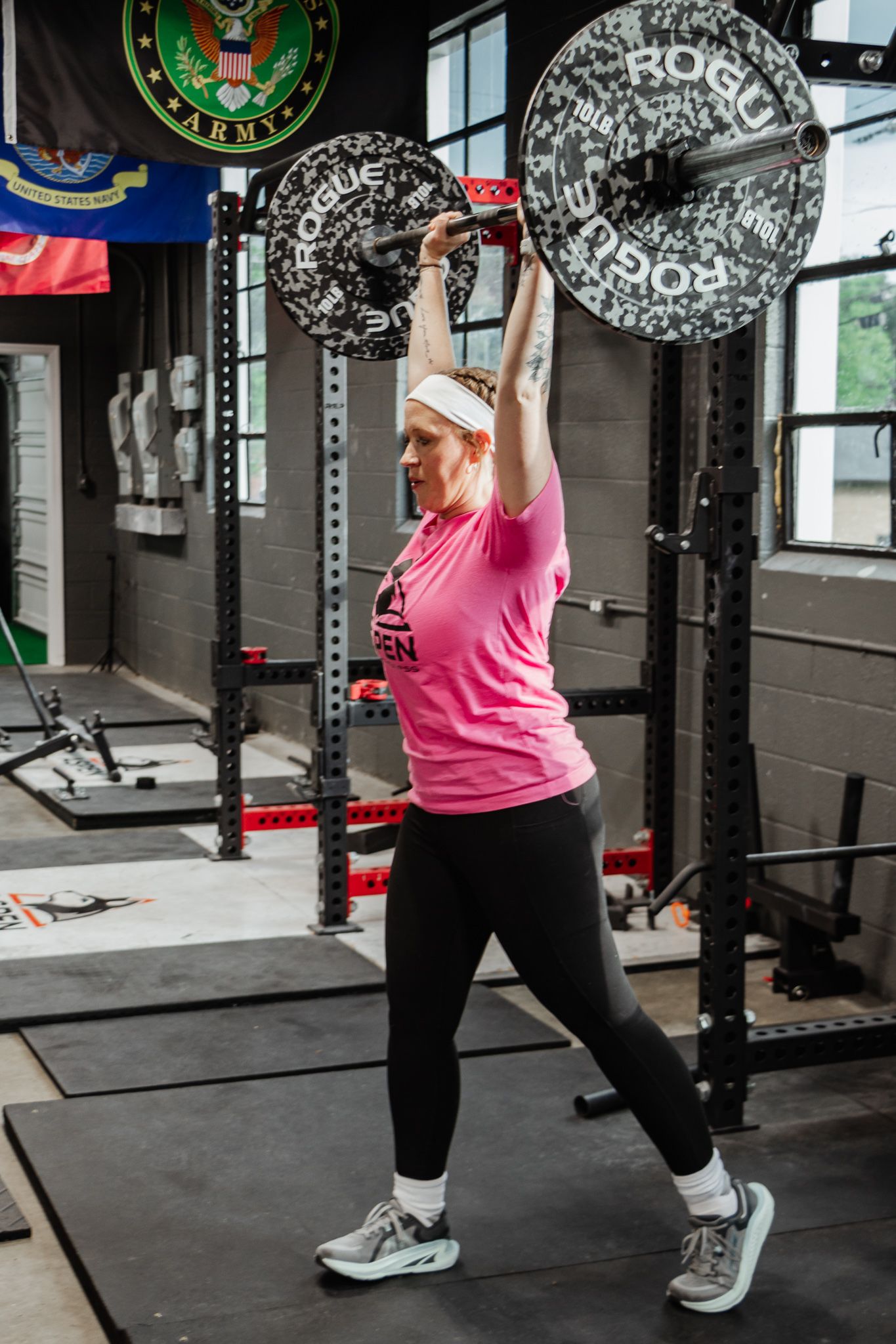 Woman in a pink shirt performs a split jerk, holding a loaded barbell overhead in a strength training gym with flags on the wall.
