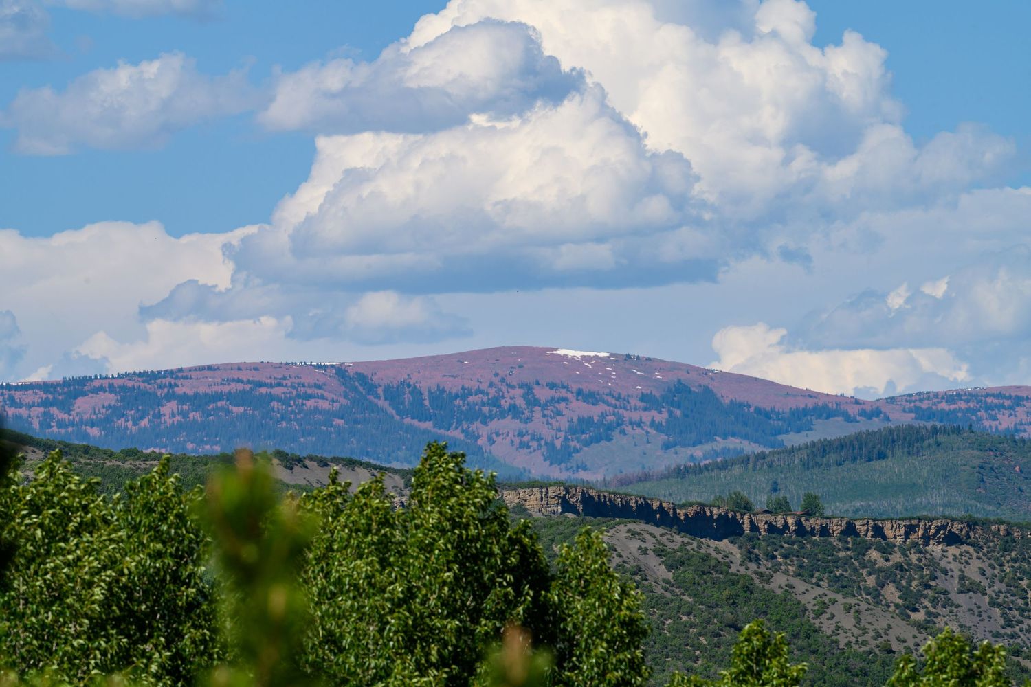 Scenic mountain landscape with pink-hued peak, forested hills, and dramatic white clouds above.