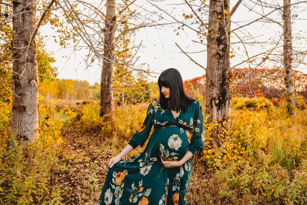 Pregnant woman in floral dress posing among autumn trees during sunset.