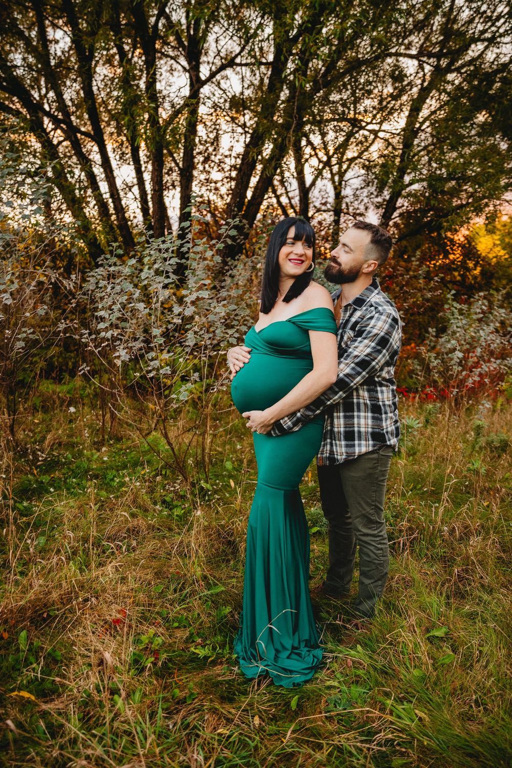 A maternity photo shoot in a forest features an emerald green dress and plaid shirt against autumn foliage.