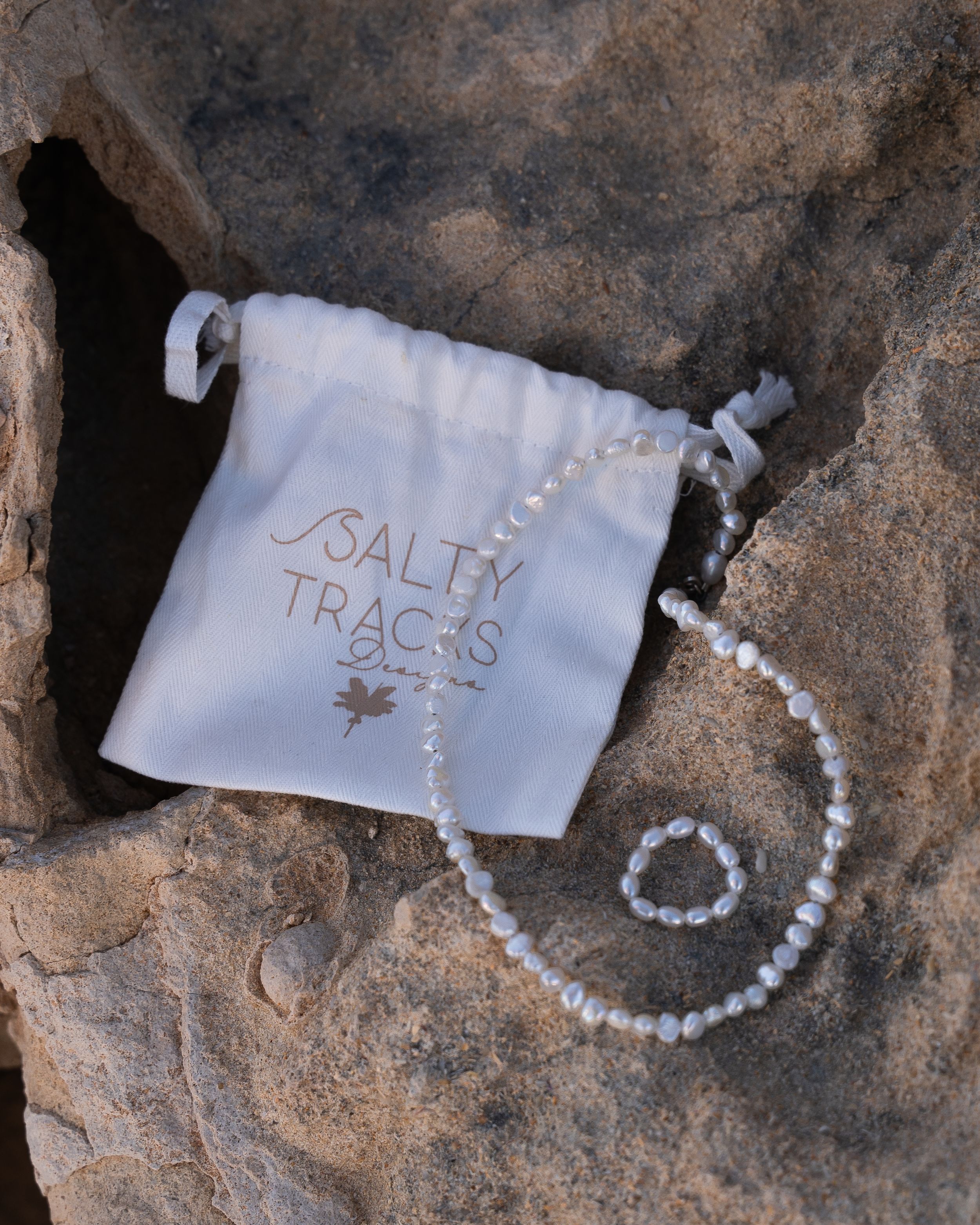 White drawstring bag labeled 'Salty Tracks' with pearl necklace on rocky surface.