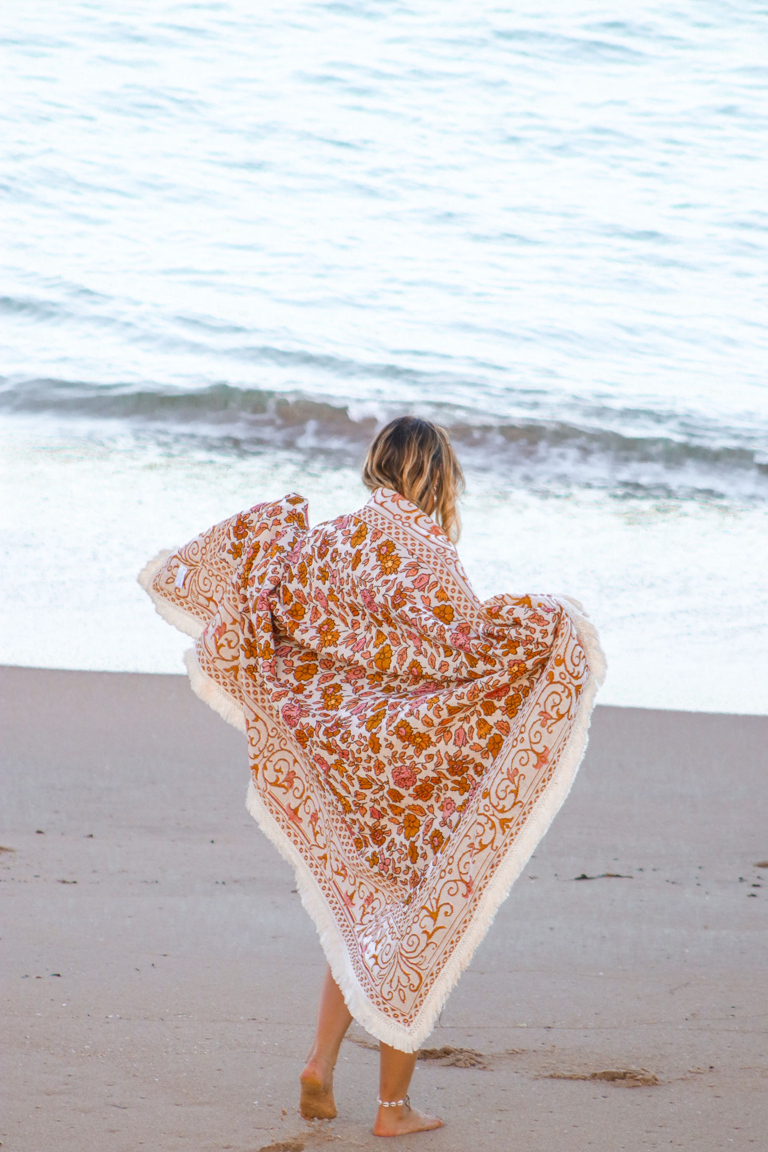 Person wrapped in patterned orange beach blanket standing by ocean waves.