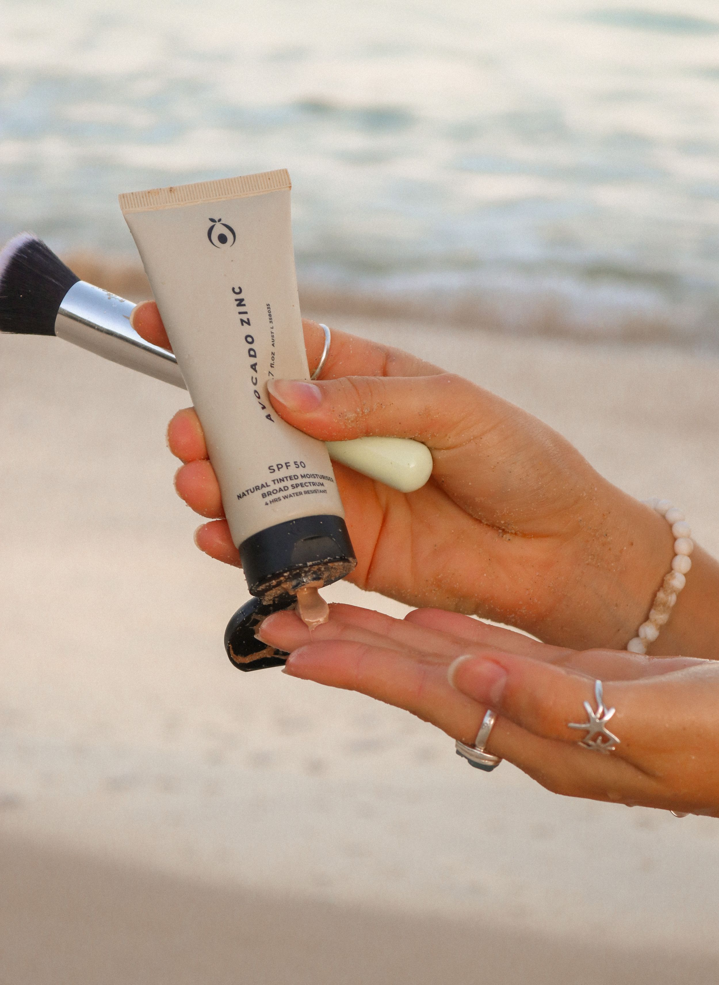 Hands holding skincare product tube against beach background with jewelry details.