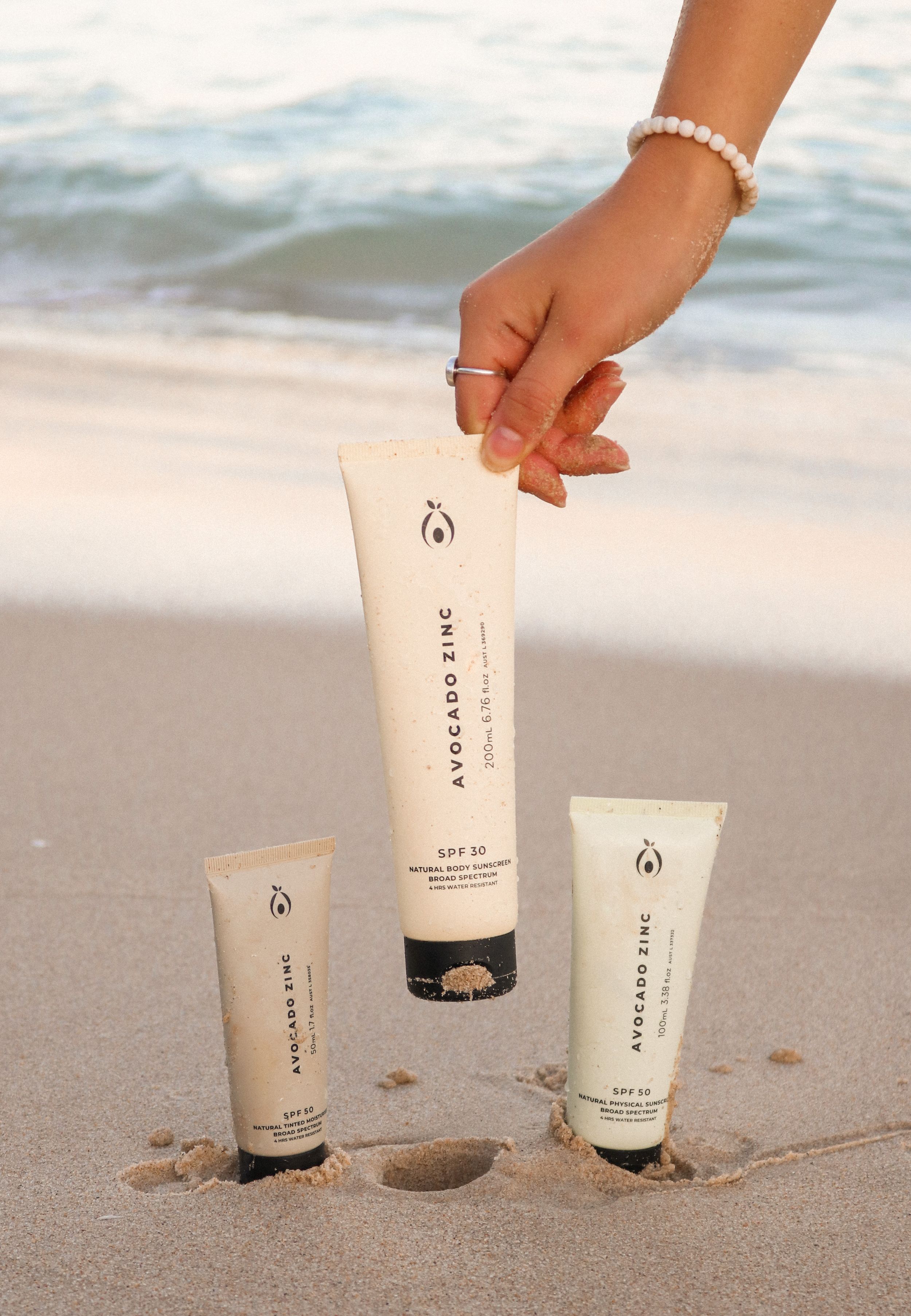 Three white cosmetic tubes arranged on sandy beach with ocean waves in background and hand reaching to grab one.