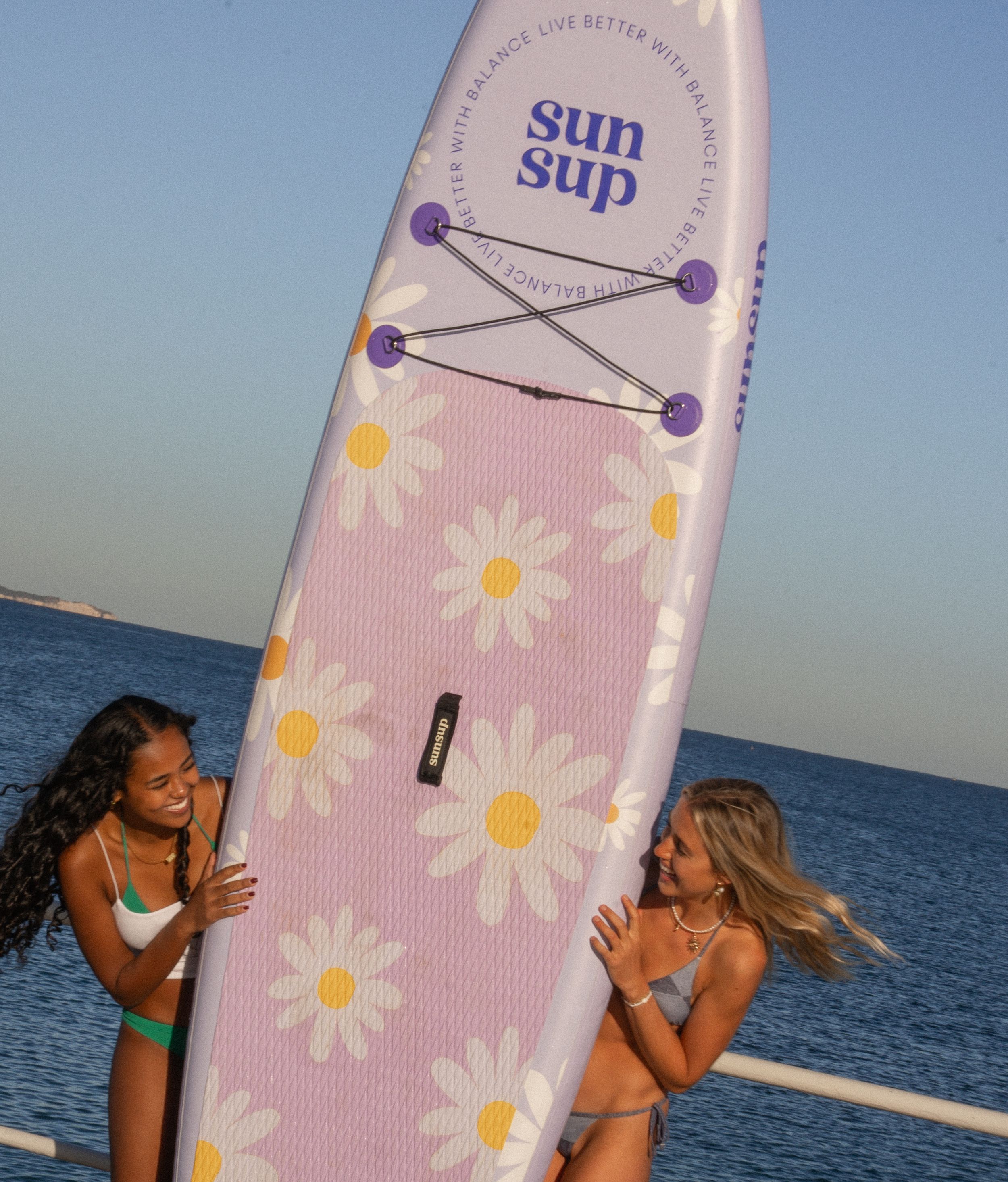 Two surfers share a playful moment on a pink floral surfboard against an ocean backdrop.