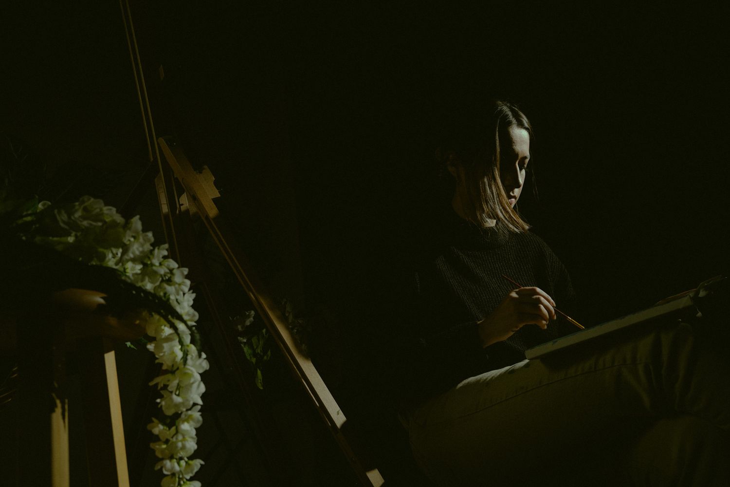 A person in dark attire prepares to paint in moody atmospheric lighting with floral decorations nearby.