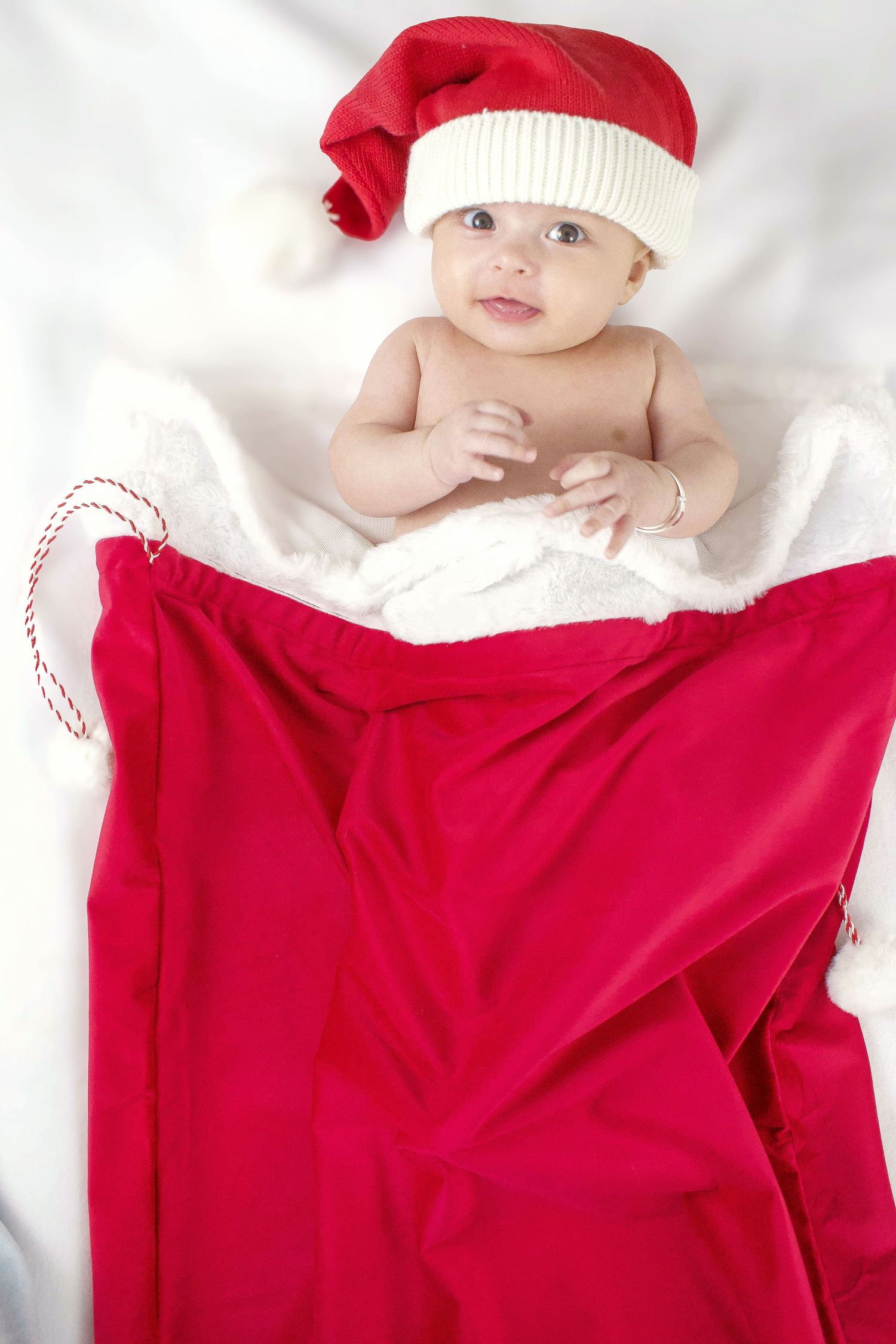 Baby wearing red Santa hat and blanket lying on white bedding during Christmas photoshoot.