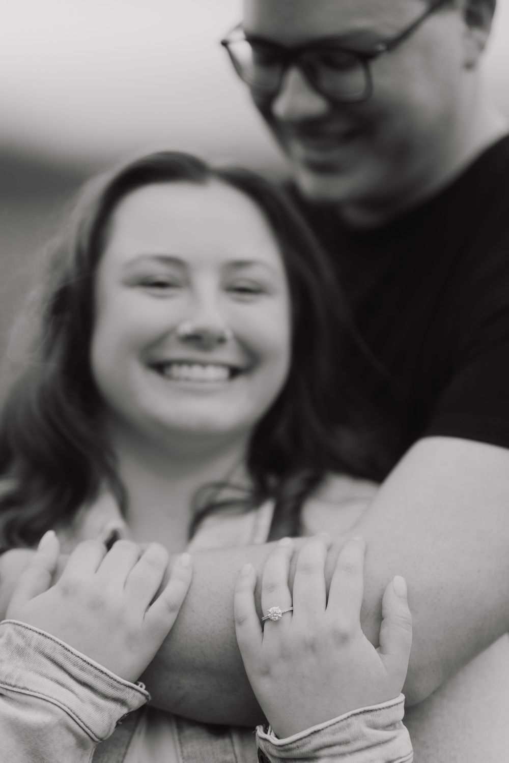 A black and white close-up of a couple embracing shows their hands with wedding rings.