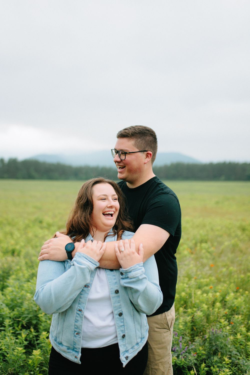 Happy couple shares joyful moment together while embracing in blooming meadow with mountains in background.