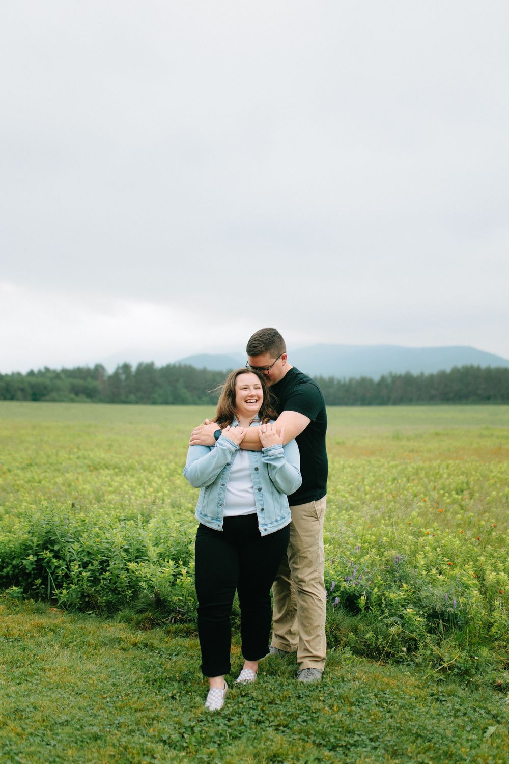 A couple embraces in a scenic meadow with rolling hills and green fields in the background on an overcast day.