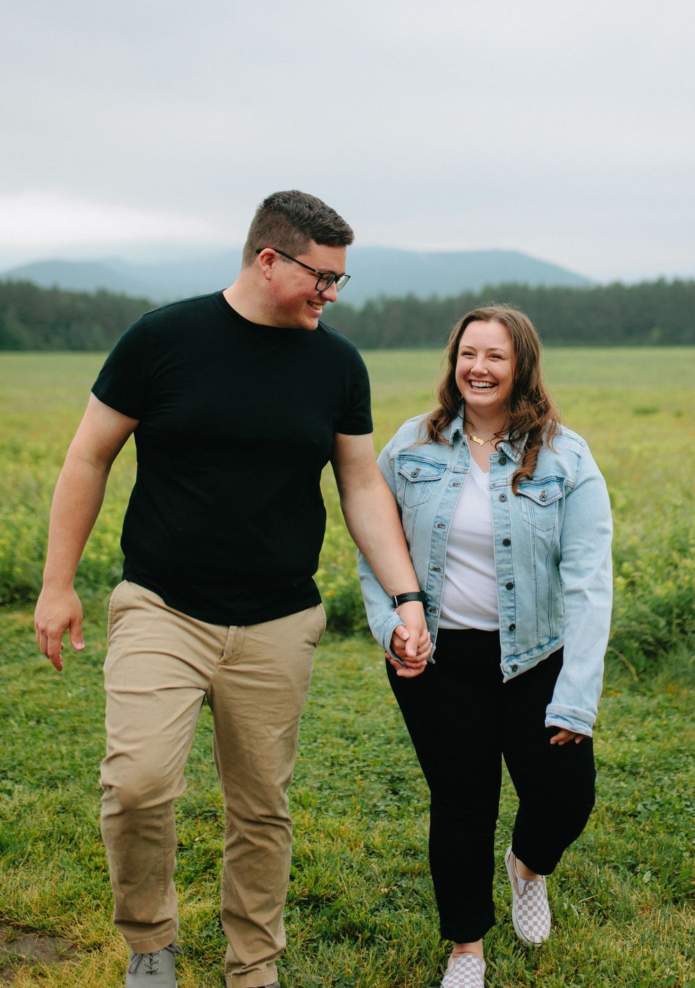 A couple wearing casual clothing holds hands while walking together in a green meadow with mountains in the background.