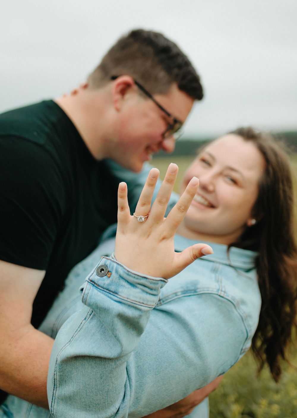 A newly engaged couple embraces outdoors while showing off an engagement ring in natural lighting.