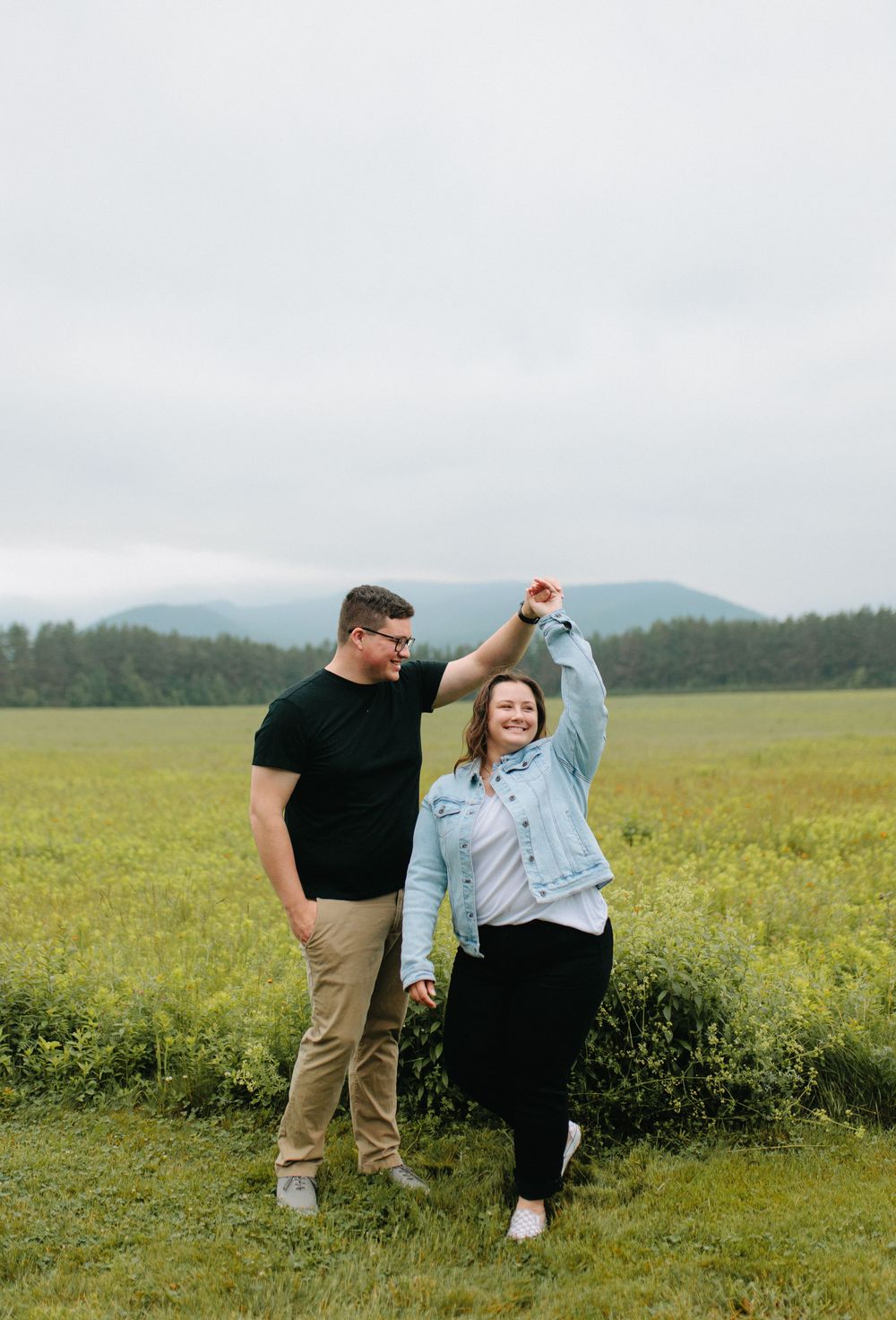 A couple dances together in a sunny meadow with mountains in the background during a romantic photoshoot.
