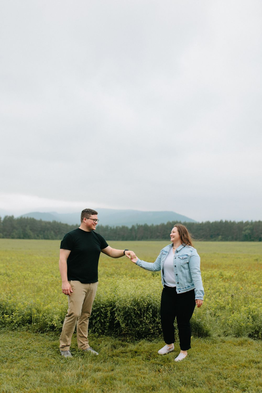 A couple holds hands in a scenic field with rolling hills and forest in the background on an overcast day.